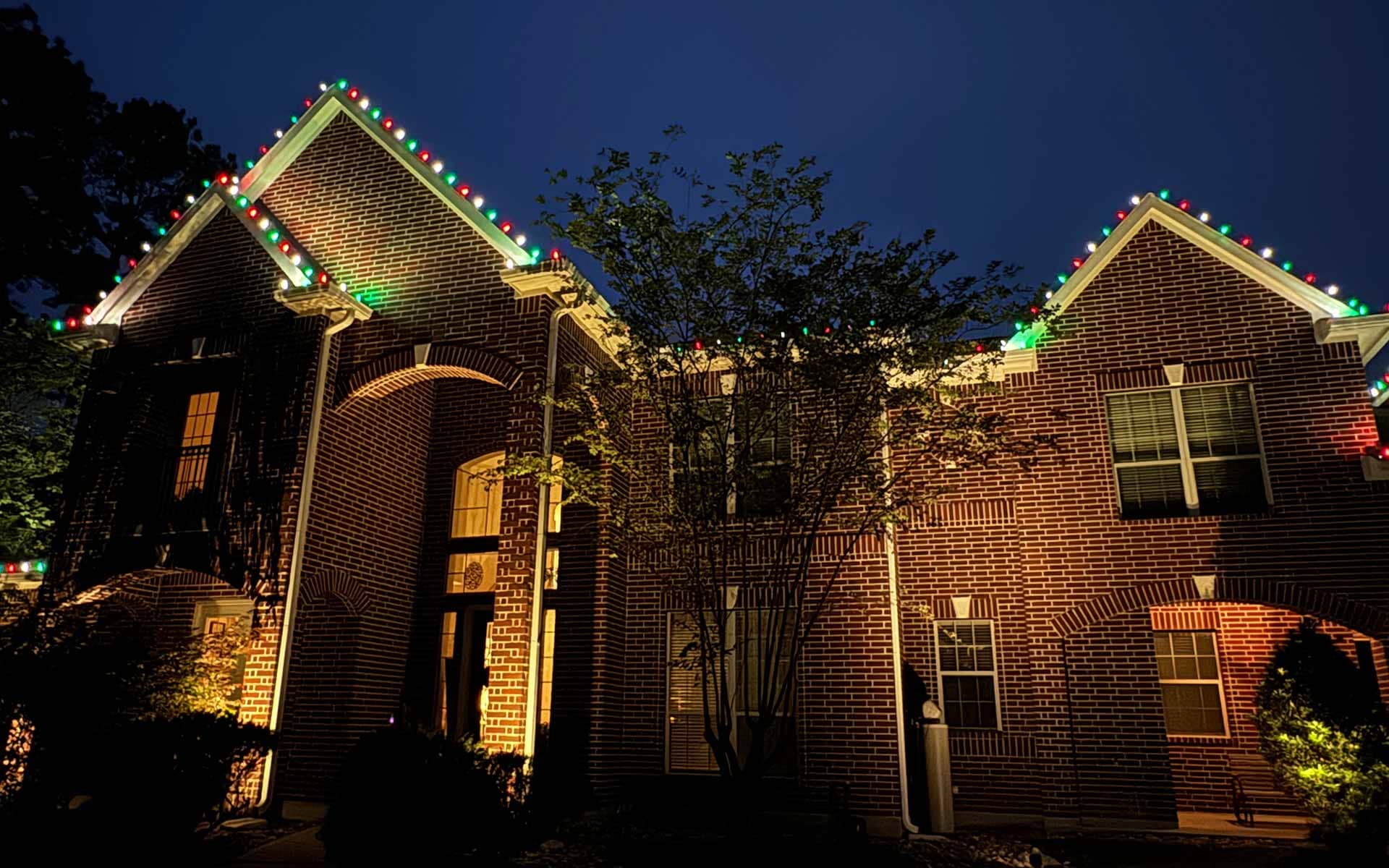 Red, green, and white Christmas lights lining the rooflines of a two-story brick home at dusk in North Houston